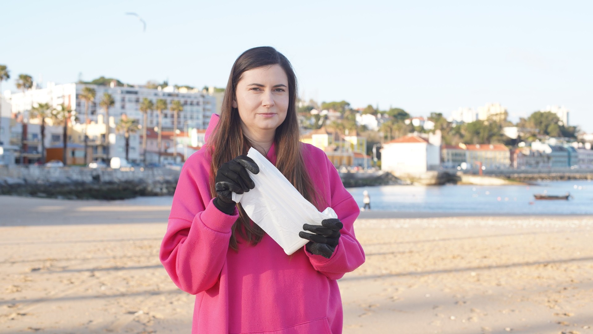 Girl in pink sweatshirt tearing off white plastic garbage bag on beach. Soft focus